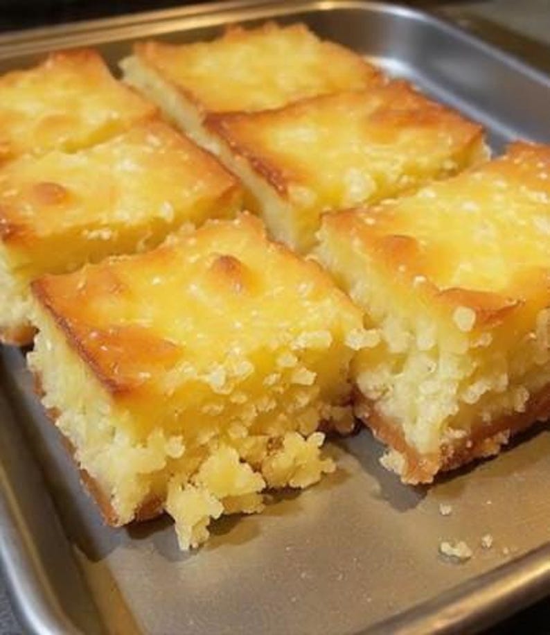 May include: A tray of golden-brown, square-cut baked goods. The tops have a glossy, caramelized appearance, and the edges are slightly darker. The texture appears moist and crumbly, suggesting a dessert like a bar or cake.