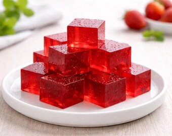 May include: A stack of translucent, bright red gelatin cubes arranged in a pyramid shape on a white plate. Each square piece has a smooth, glossy surface with small water droplets visible on the edges, reflecting light through the vibrant red color.