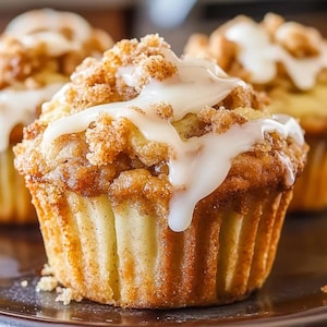 May include: Three golden-brown muffins with a pleated paper liner, topped with a textured cinnamon brown sugar crumble and drizzled with a thick, opaque white icing glaze. The muffins have a light tan interior and a baked, caramelized exterior.