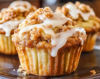 May include: Three golden-brown muffins with a pleated paper liner, topped with a textured cinnamon brown sugar crumble and drizzled with a thick, opaque white icing glaze. The muffins have a light tan interior and a baked, caramelized exterior.