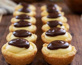 May include: A close-up of a wooden surface displays rows of mini tarts. Each tart has a golden-brown, baked crust filled with a creamy, pale yellow custard. A generous topping of dark chocolate covers the custard, creating a visually appealing dessert.