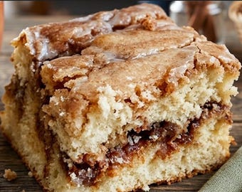 May include: A square slice of coffee cake with a light brown, crumbly topping and a layer of cinnamon filling. The cake has a golden-brown crust and is drizzled with a white glaze. The image shows the cake's texture and layers.