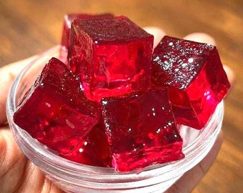May include: A clear glass bowl filled with vibrant red, cube-shaped gelatin desserts. The gelatin appears glossy and slightly translucent, with a glistening surface. The cubes are stacked, creating a visually appealing arrangement.
