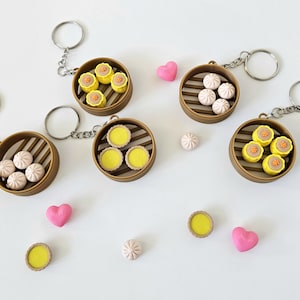 May include: Keychains featuring miniature dim sum, including dumplings and egg tarts, presented in bamboo steamer baskets. The keychains have silver rings and chains. Pink heart-shaped decorations and individual dim sum pieces are scattered around the keychains.