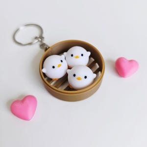 May include: A keychain featuring three white, round chick-shaped charms with black eyes and orange beaks, nestled in a brown bamboo steamer. Two pink heart-shaped objects are also present on a white surface.