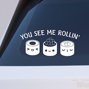 以下が含まれることがあります： 白いビニール製の車のステッカー。笑顔の3つの寿司ロールが描かれています。「YOU SEE ME ROLLIN'」の文字が寿司の上にあります。ステッカーは車の窓に貼られています。
