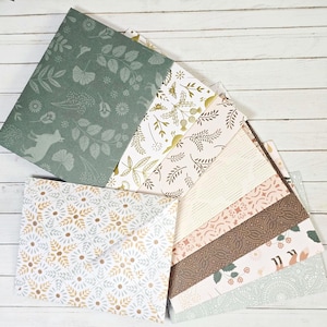 May include: A collection of patterned paper sheets and envelopes. The designs include floral, leaf, and geometric patterns in shades of green, gold, brown, and white. The papers are arranged on a white wooden surface.