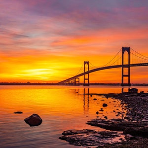 May include: A silhouette of a bridge against a vibrant orange and pink sunset over a calm body of water. The bridge has a suspension design with multiple towers and cables.