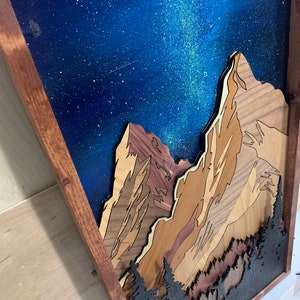 Puede incluir: Una pieza de arte de pared de madera que representa una cordillera con un cielo nocturno estrellado. Las monta&ntilde;as est&aacute;n hechas de diferentes tipos de madera y est&aacute;n superpuestas para crear un efecto 3D. El cielo est&aacute; pintado con un fondo azul y estrellas blancas, con una V&iacute;a L&aacute;ctea verde y blanca visible.