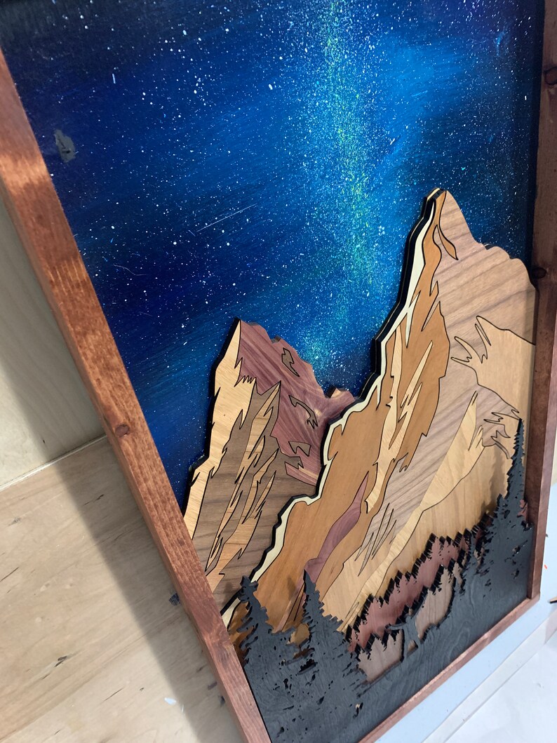 Puede incluir: Una pieza de arte de pared de madera que representa una cordillera con un cielo nocturno estrellado. Las monta&ntilde;as est&aacute;n hechas de diferentes tipos de madera y est&aacute;n superpuestas para crear un efecto 3D. El cielo est&aacute; pintado con un fondo azul y estrellas blancas, con una V&iacute;a L&aacute;ctea verde y blanca visible.