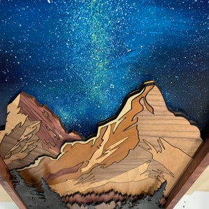 Puede incluir: Una pieza de arte de pared de madera que representa una cordillera con un cielo nocturno estrellado. Las monta&ntilde;as est&aacute;n hechas de diferentes tipos de madera, creando un efecto de capas. El cielo est&aacute; pintado con un fondo azul oscuro y estrellas blancas, con una V&iacute;a L&aacute;ctea verde y blanca visible.