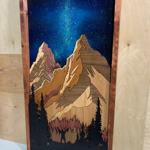Puede incluir: Una pieza de arte de pared de madera que representa una cordillera bajo un cielo nocturno estrellado. Las monta&ntilde;as est&aacute;n hechas de diferentes tipos de madera, creando un efecto de capas. El cielo est&aacute; pintado de azul con estrellas blancas y una galaxia de la V&iacute;a L&aacute;ctea. Una silueta de una persona de pie frente a un bosque es visible en la parte inferior de la pieza.