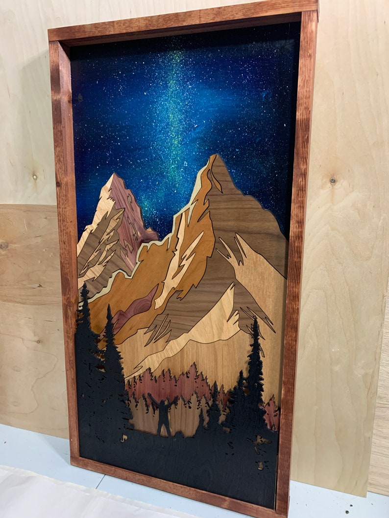 Puede incluir: Una pieza de arte de pared de madera que representa una cordillera bajo un cielo nocturno estrellado. Las monta&ntilde;as est&aacute;n hechas de diferentes tipos de madera, creando un efecto de capas. El cielo est&aacute; pintado de azul con estrellas blancas y una galaxia de la V&iacute;a L&aacute;ctea. Una silueta de una persona de pie frente a un bosque es visible en la parte inferior de la pieza.