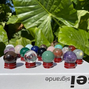 Könnte beinhalten: Eine Sammlung polierter Edelstein-Kugeln in verschiedenen Farben, darunter Amethyst, Rosenquarz und Malachit, die auf kleinen, roten Holzständern ausgestellt sind. Die Kugeln sind auf einer weißen Oberfläche angeordnet, mit üppigem grünem Laub im Hintergrund.