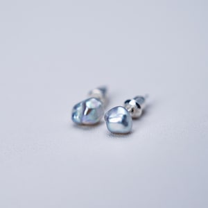 May include: A pair of light blue pearl stud earrings. The earrings have a unique, irregular shape and a subtle iridescent sheen. The posts are silver. The earrings are displayed on a white surface.