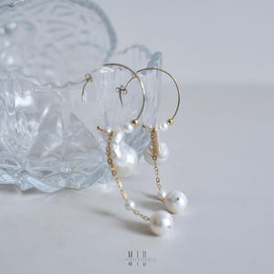 May include: Elegant gold hoop earrings with dangling pearls. The earrings feature a delicate chain and a mix of large and small pearls. The design includes a large baroque pearl and several smaller, round pearls. The earrings are displayed near a clear glass dish.