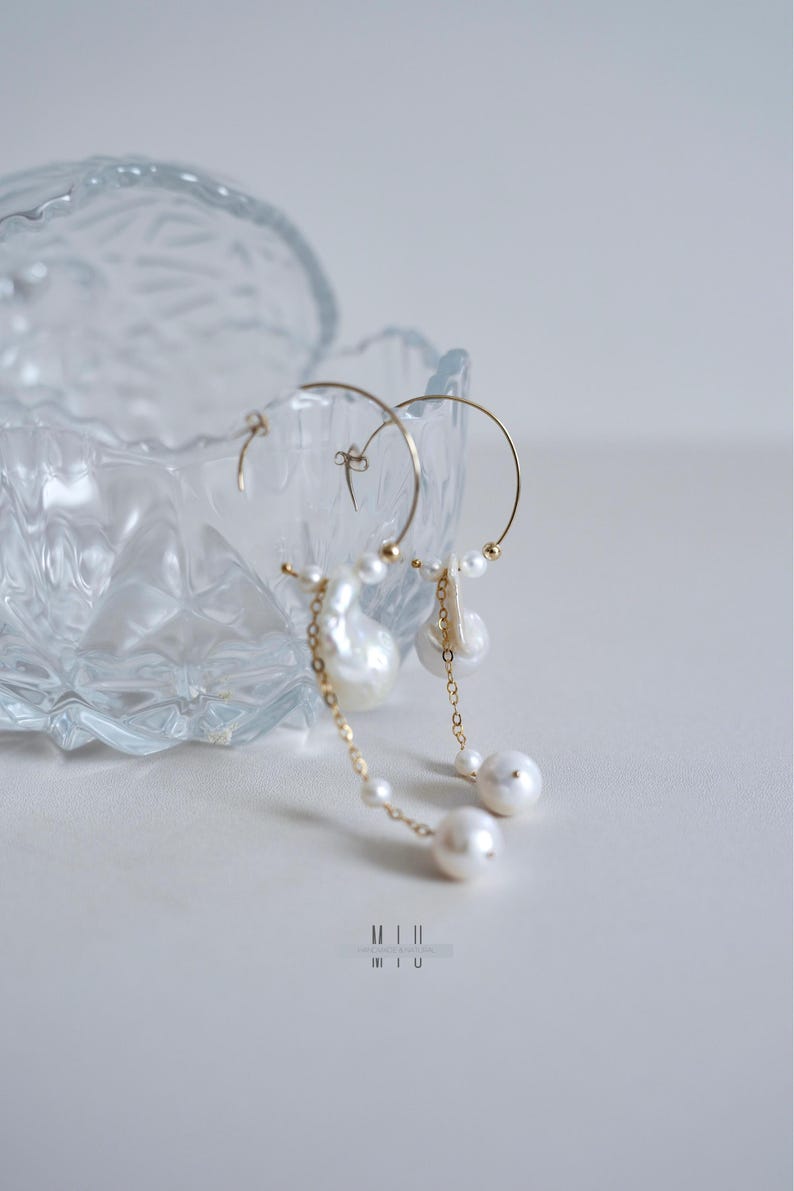May include: Elegant gold hoop earrings with dangling pearls. The earrings feature a delicate chain and a mix of large and small pearls. The design includes a large baroque pearl and several smaller, round pearls. The earrings are displayed near a clear glass dish.