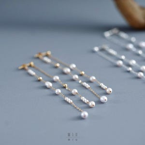 Pu&ograve; includere: Eleganti orecchini dorati con perle bianche pendenti e piccole perline dorate. Gli orecchini sono disposti su una superficie grigia, mostrando il loro design delicato. Un accessorio elegante.