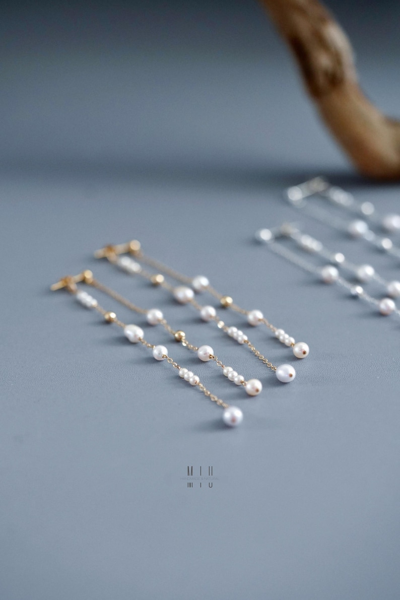 Pu&ograve; includere: Eleganti orecchini dorati con perle bianche pendenti e piccole perline dorate. Gli orecchini sono disposti su una superficie grigia, mostrando il loro design delicato. Un accessorio elegante.