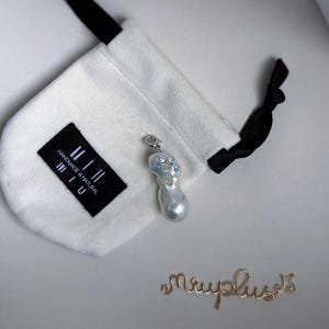 Op de afbeelding: Een barokke parel hanger met een zilveren beugel en kleine heldere stenen, naast een witte trekkoordtas met een zwart label met de tekst "handmade & natural miu". Een gouden draadscript spelt "miuplus".