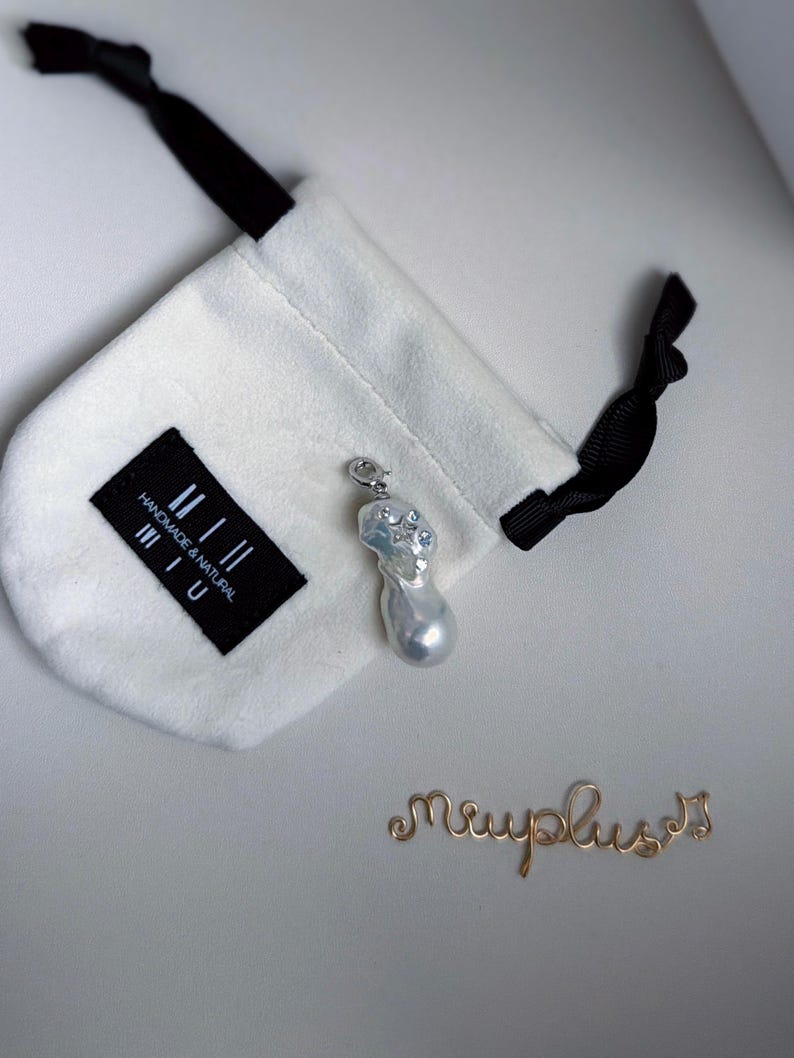 Op de afbeelding: Een barokke parel hanger met een zilveren beugel en kleine heldere stenen, naast een witte trekkoordtas met een zwart label met de tekst "handmade & natural miu". Een gouden draadscript spelt "miuplus".