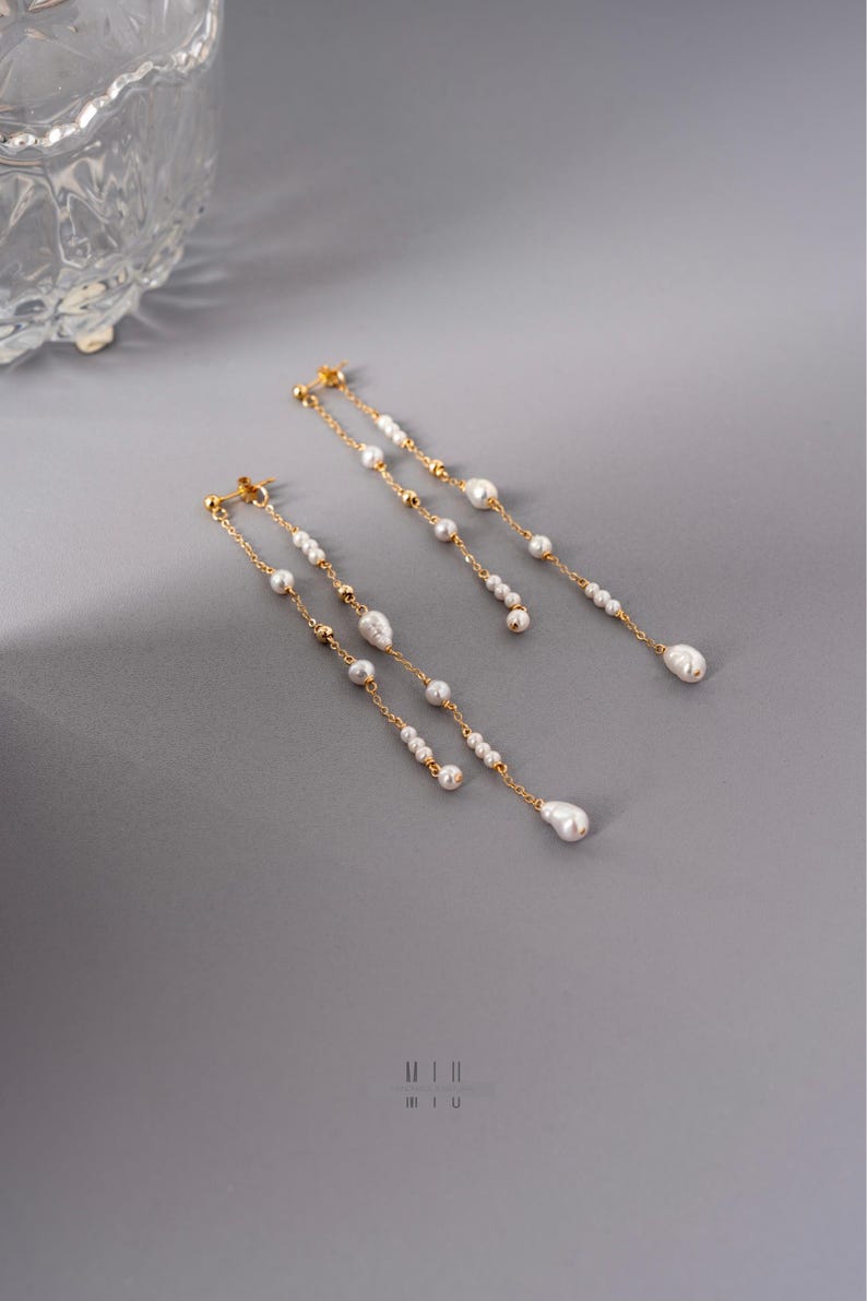 Pu&ograve; includere: Eleganti orecchini dorati con perle bianche pendenti. Gli orecchini presentano un delicato design a catena con molteplici piccole perle e una perla pi&ugrave; grande a forma di goccia all'estremit&agrave;. Su sfondo grigio.