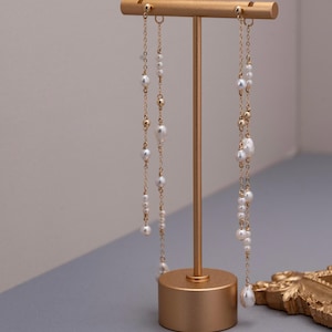 Pu&ograve; includere: Un espositore per orecchini color oro che tiene due paia di orecchini pendenti. Gli orecchini presentano perle bianche e perline color oro infilate su delicate catene. L'espositore &egrave; cilindrico con una barra orizzontale nella parte superiore.