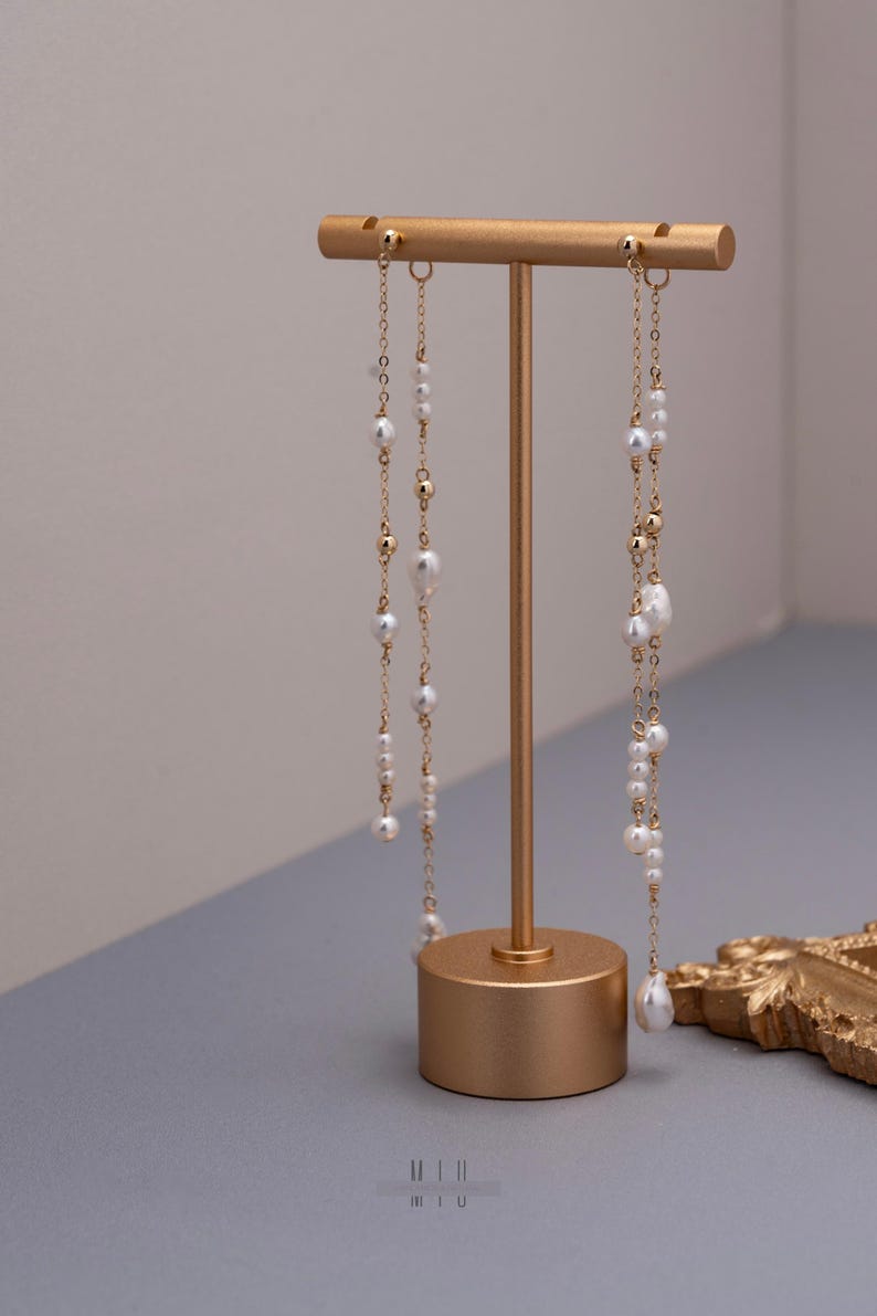 Pu&ograve; includere: Un espositore per orecchini color oro che tiene due paia di orecchini pendenti. Gli orecchini presentano perle bianche e perline color oro infilate su delicate catene. L'espositore &egrave; cilindrico con una barra orizzontale nella parte superiore.