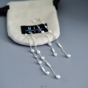 Pu&ograve; includere: Orecchini in argento con perle bianche pendenti. Sono presentati vicino a un piccolo sacchetto color crema con un'etichetta nera. L'immagine, con messa a fuoco morbida, mette in risalto i gioielli.