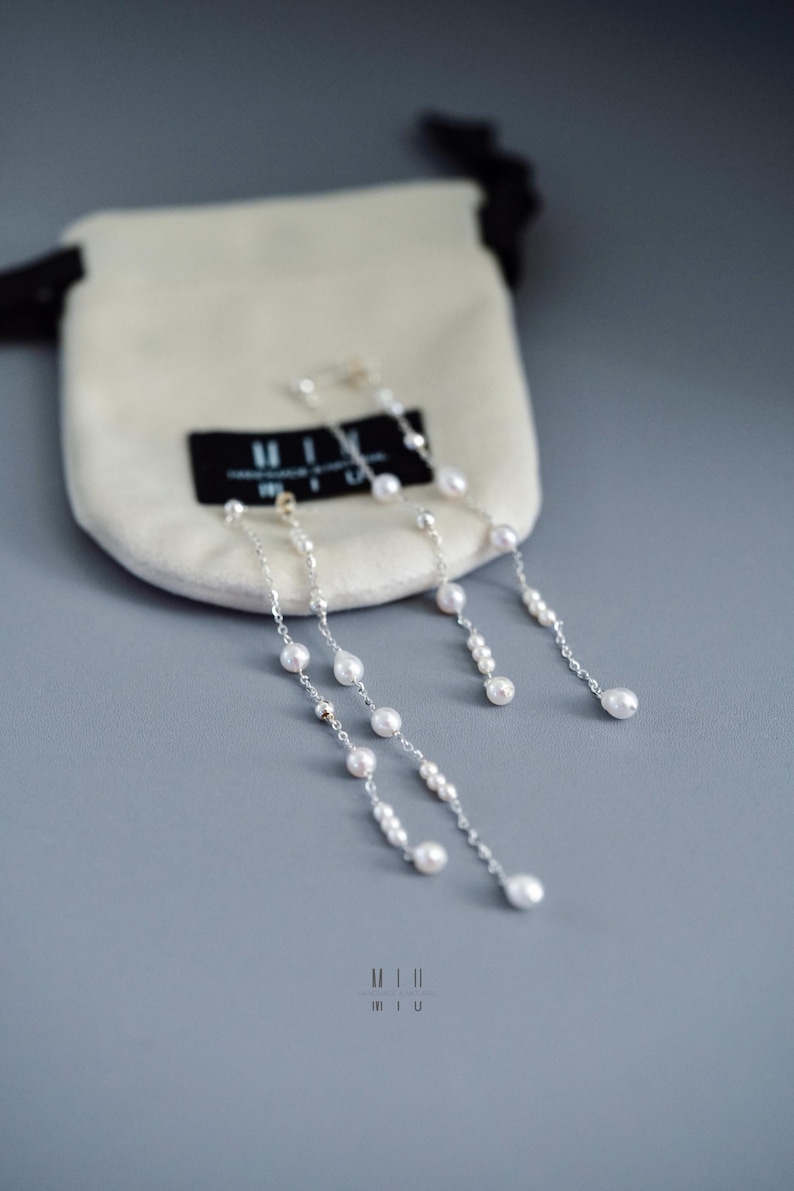 Pu&ograve; includere: Orecchini in argento con perle bianche pendenti. Sono presentati vicino a un piccolo sacchetto color crema con un'etichetta nera. L'immagine, con messa a fuoco morbida, mette in risalto i gioielli.