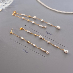 Pu&ograve; includere: Eleganti orecchini color oro con perle bianche pendenti. Un paio &egrave; lungo 8 cm, l'altro 9 cm. Gli orecchini presentano piccole perline dorate e una perla pi&ugrave; grande a forma di goccia all'estremit&agrave;. I gioielli sono esposti su una superficie grigia.