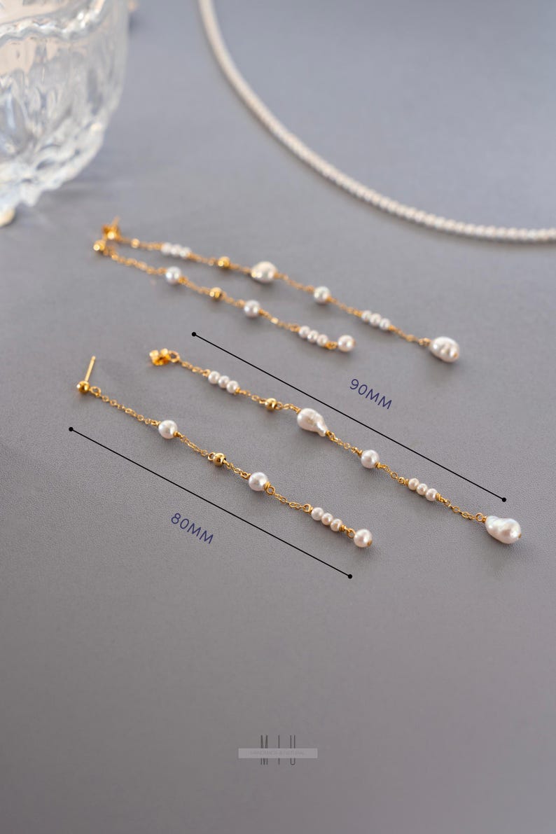 Pu&ograve; includere: Eleganti orecchini color oro con perle bianche pendenti. Un paio &egrave; lungo 8 cm, l'altro 9 cm. Gli orecchini presentano piccole perline dorate e una perla pi&ugrave; grande a forma di goccia all'estremit&agrave;. I gioielli sono esposti su una superficie grigia.