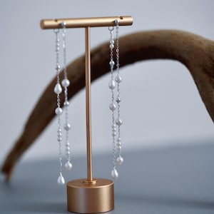 Pu&ograve; includere: Eleganti orecchini in argento con perle bianche pendenti, esposti su un supporto color oro. Gli orecchini presentano un delicato design a catena con perle luminose di varie dimensioni. Il supporto ha un'estetica semplice e moderna.