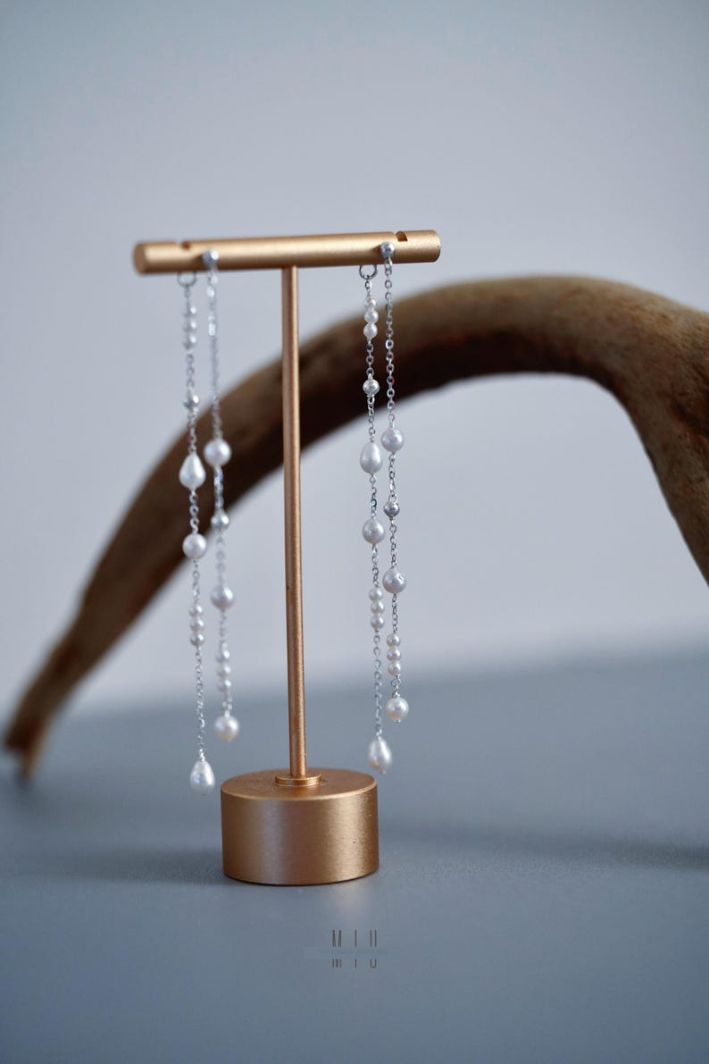 Pu&ograve; includere: Eleganti orecchini in argento con perle bianche pendenti, esposti su un supporto color oro. Gli orecchini presentano un delicato design a catena con perle luminose di varie dimensioni. Il supporto ha un'estetica semplice e moderna.
