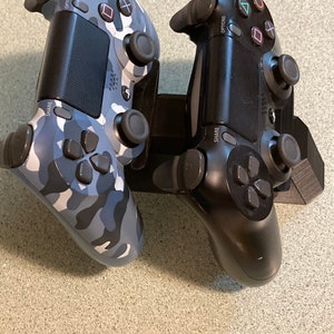May include: Two black PlayStation 4 controllers, one with a camouflage design, are mounted on a black stand. The controllers have the words "Share" and "Options" printed on them.