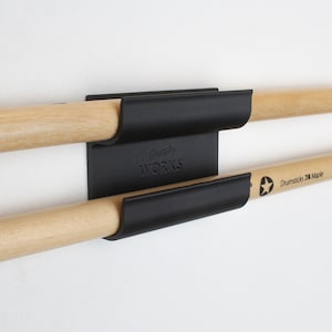May include: A black metal drumstick holder mounted on a white wall, holding two wooden drumsticks. The drumsticks are light brown with the text "Drumsticks 7A Maple" printed on one. The holder has the text "Gravity WORKS" on it.