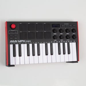 May include: A black and red AKAI MPK Mini MIDI keyboard with white keys. The keyboard has a red trim and a red button. It features several knobs, pads, and a display screen. The product is designed for music production.