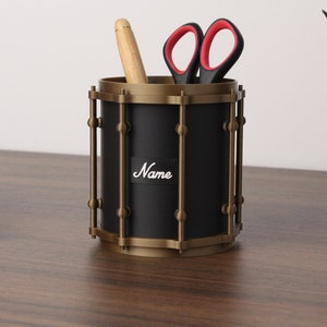 Pode incluir: Um organizador de mesa preto e dourado em forma de tambor com o nome "Name" na frente. Ele contém uma caneta de madeira e tesouras pretas com cabos vermelhos. Um teclado pequeno e uma planta em vaso estão no fundo.