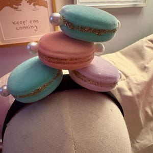 May include: A black headband adorned with four plush, pastel-colored macaron-shaped decorations. Each macaron is a different color: teal, pink, and lavender, with a gold glitter band and a pearl accent. The macarons are stacked on top of each other.