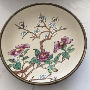 May include: A round ceramic bowl with a cream-colored base and a metallic rim. The interior features a hand-painted design of brown branches with green leaves, small white blossoms, and three large, vibrant purple flowers with yellow centers.