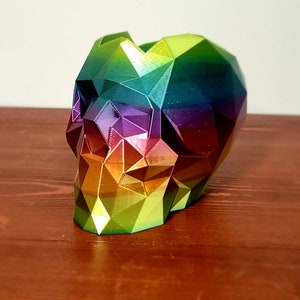 Puede incluir: Un macetero de calavera iridiscente arcoíris hecho de plástico impreso en 3D. La calavera es un diseño geométrico con una superficie facetada.