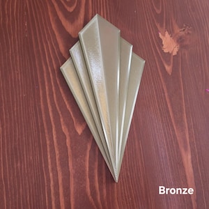 Puede incluir: Un adorno decorativo de pared de color bronce que presenta una disposición en abanico de cinco segmentos alargados y puntiagudos que convergen en la parte inferior. El objeto tiene un acabado metálico liso y la palabra Bronze en la parte inferior.