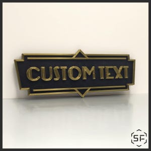 Puede incluir: Un letrero rectangular negro con borde dorado y las palabras "CUSTOM TEXT" en letras doradas. El letrero tiene un borde decorativo con formas geométricas en las esquinas. El letrero está sobre una superficie blanca.