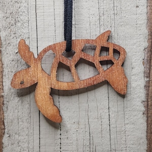 May include: A wooden turtle ornament with a dark cord for hanging. The turtle is cut out with a detailed design, showcasing the natural wood grain. The background is a weathered, light-colored wooden surface.