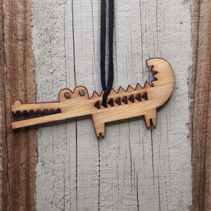 May include: A wooden alligator ornament with a dark blue string for hanging. The alligator is light brown with etched details, including teeth and scales. The ornament is hanging against a weathered, light-colored wooden background.
