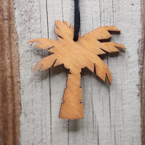May include: A wooden palm tree ornament with a dark blue string for hanging. The palm tree is light brown with detailed cutouts of the leaves and trunk. The background is a weathered, light-colored wooden surface.