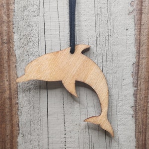May include: A wooden dolphin pendant with a black cord. The dolphin is cut from light-colored wood, showcasing the natural grain. The pendant is suspended against a rustic, weathered wood background.