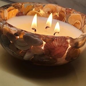 May include: A decorative candle with three lit wicks, encased in a clear bowl. The bowl is adorned with seashells of various sizes and colors, creating a coastal theme. The candle is white, and the flames are bright.