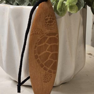 May include: A wooden surfboard-shaped ornament with a detailed turtle carving. The ornament is light brown with a black cord for hanging. The turtle design is etched into the wood, showcasing the texture and detail of the carving.