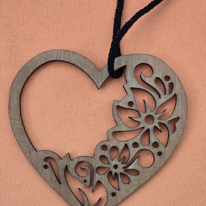 May include: Wooden heart-shaped ornament with a floral design cut into the right side. The ornament is a light brown color and has a black cord for hanging. The background is a solid peach color.
